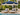 A navy blue patio umbrella with cream fringe trim stands between two cream chaise lounge chairs on a poolside patio. The scene is surrounded by tropical greenery, palm trees, and a modern house exterior.